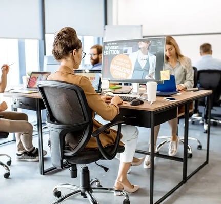 Une femme est assise à un bureau, concentrée sur l'écran de son ordinateur, entourée de collègues travaillant dans un environnement de bureau lumineux.
