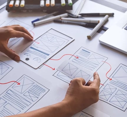 Les mains esquissent des wireframes sur papier, les reliant par des lignes rouges, à côté de marqueurs et d'un ordinateur portable, illustrant un processus de conception.