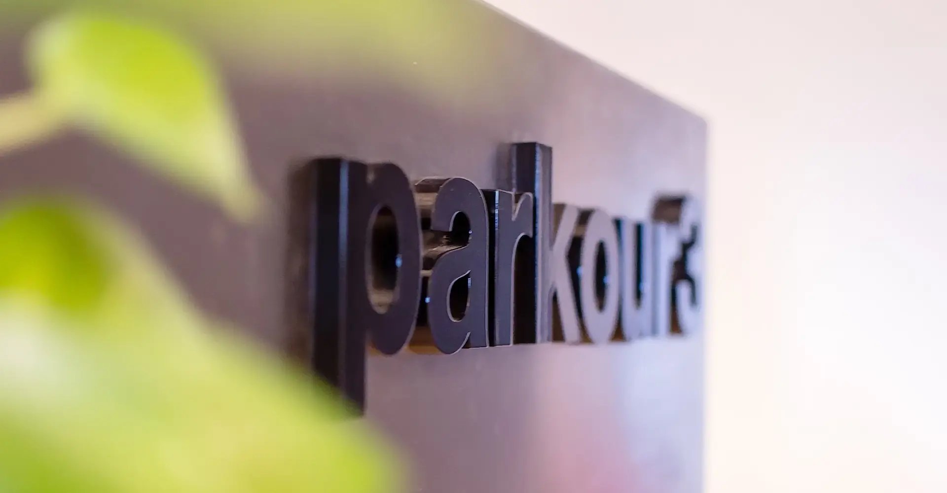 Logo noir de Parkour3 en trois dimensions plaqué sur une surface lisse, partiellement masqué par une plante verte.