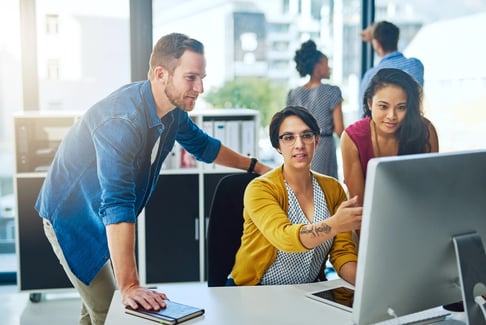 Trois personnes sont réunies autour d'un écran d'ordinateur suivant une formation en marketing dans un bureau moderne, travaillant ensemble, tandis que deux autres personnes collaborent à l'arrière-plan.