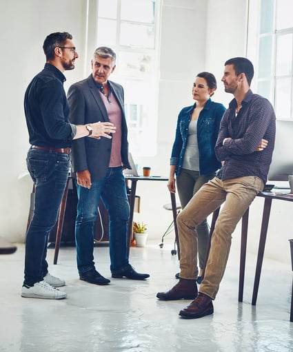 Une discussion de groupe professionnelle dans un bureau lumineux, avec quatre personnes engagées dans une conversation près d'un bureau et d'un ordinateur.