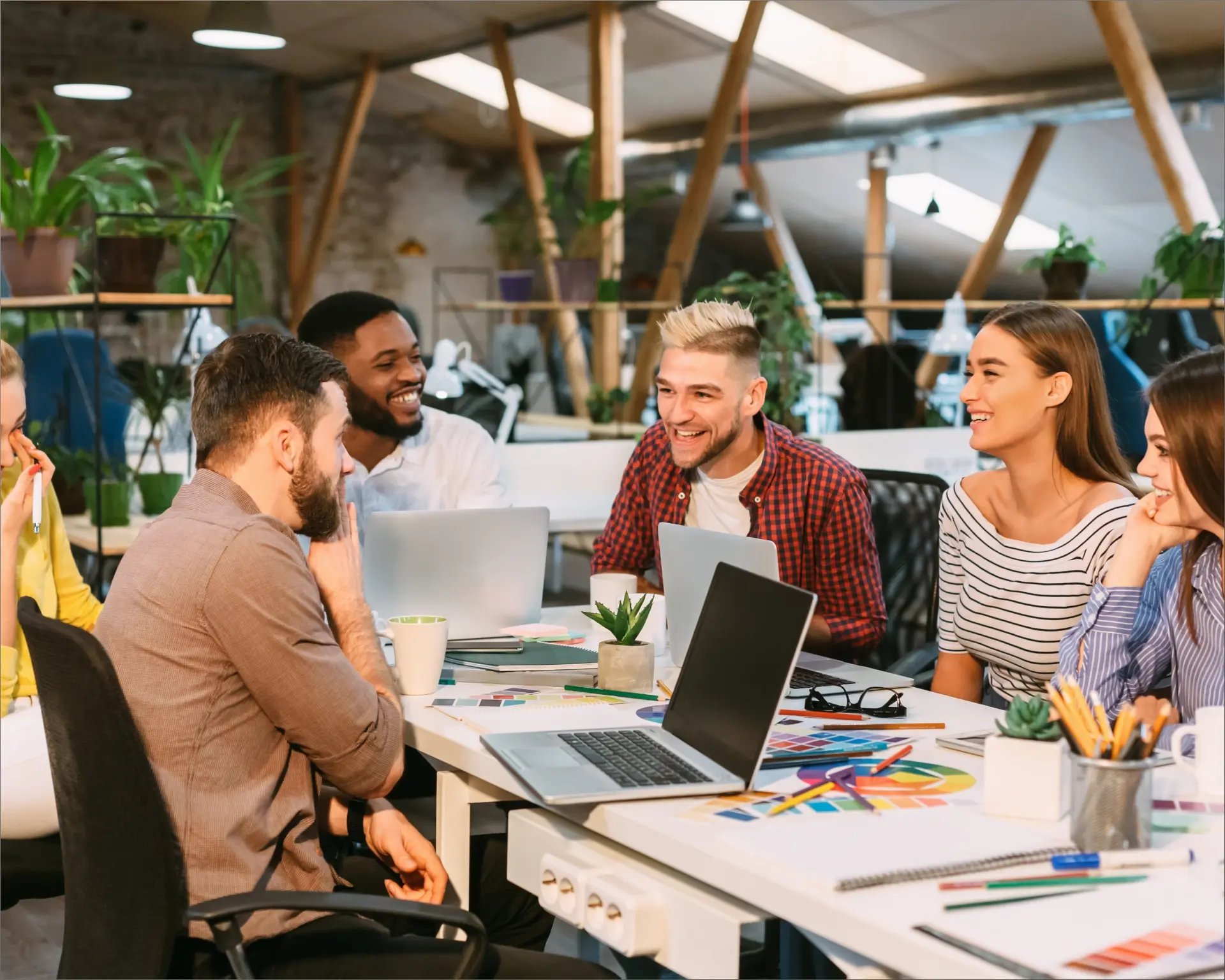 Un groupe de personnes diverses collabore de manière très amicale autour d'une table remplie d'ordinateurs portables, de cahiers et de plantes dans un espace de travail moderne et lumineux.