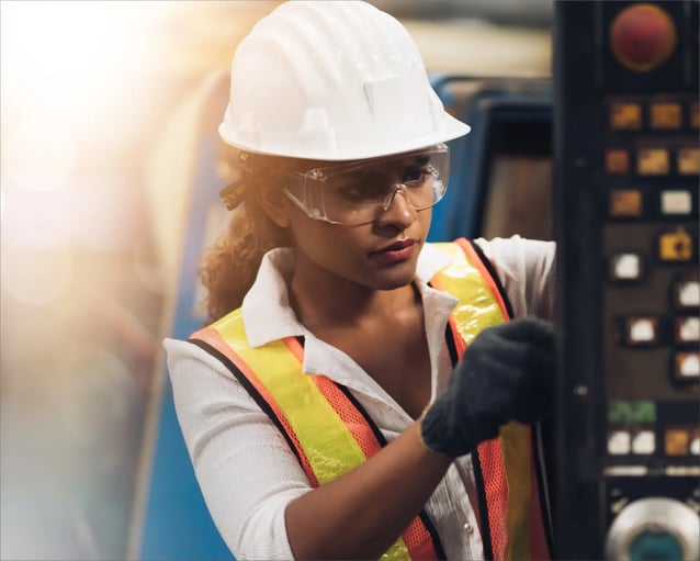 Un ouvrier portant un casque et un gilet de sécurité fait fonctionner une machine, concentré sur un panneau de commande dans un environnement industriel lumineux.