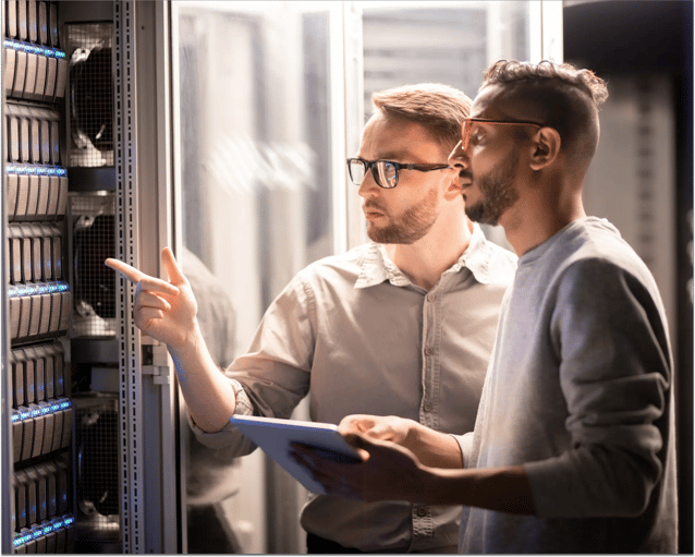 Deux personnes discutent de technologie dans une salle de serveurs, pointant du doigt des équipements tout en tenant une tablette, entourées de racks de serveurs.