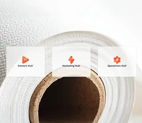Trois icônes de hub de HubSpot : Content Hub, Marketing Hub et Operations Hub sont affichées sur un fond texturé de papier blanc.