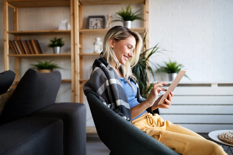 Une personne est confortablement assise dans un salon moderne, absorbée par une tablette, vêtue d'une couverture douillette et d'un pantalon jaune décontracté.