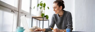 Une femme vêtue d'un pull gris étudie ou travaille à un bureau avec un ordinateur portable, entourée de plantes d'intérieur et d'un bol de snacks.
