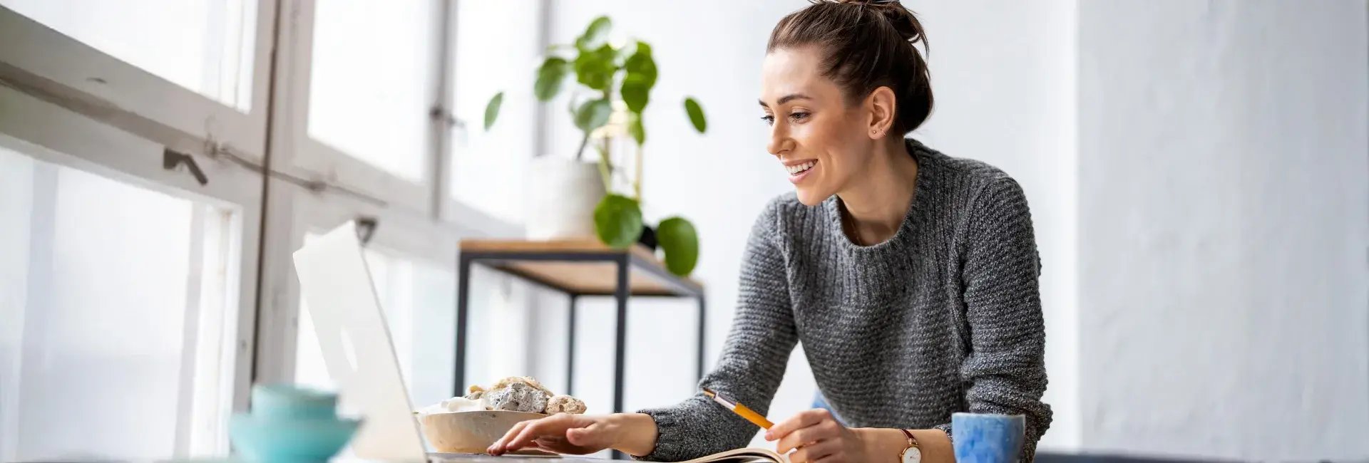 Une femme vêtue d'un pull gris étudie ou travaille à un bureau avec un ordinateur portable, entourée de plantes d'intérieur et d'un bol de snacks.