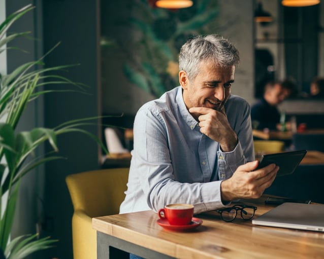 Un homme vêtu d'une chemise bleu clair est assis à une table de café, tenant une tablette, avec une tasse de café et des verres à côté de lui. Des plantes encadrent la scène.