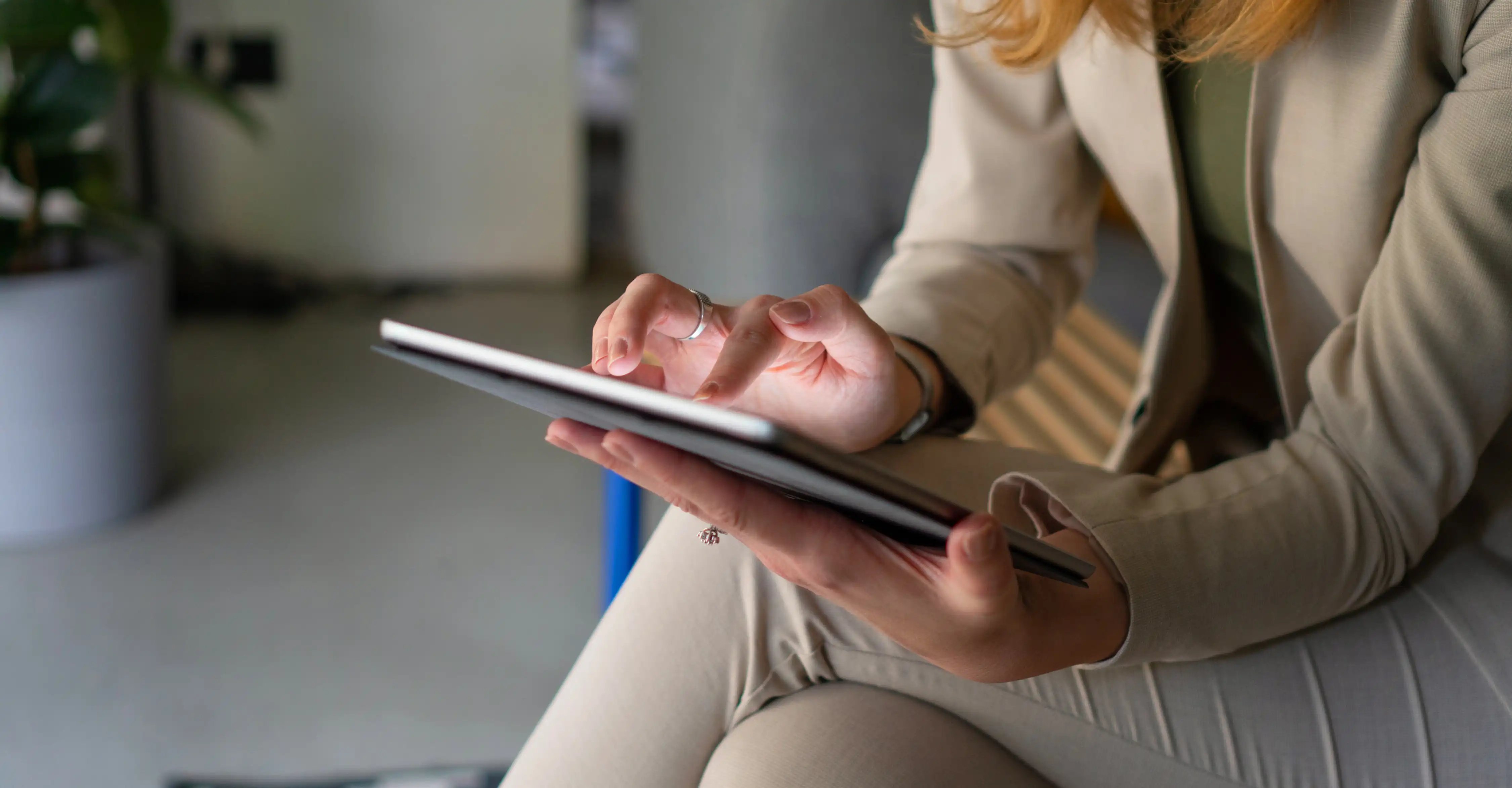 Une femme en tailleur beige utilise une tablette, tapotant l'écran avec concentration, assise dans un environnement moderne et bien éclairé.