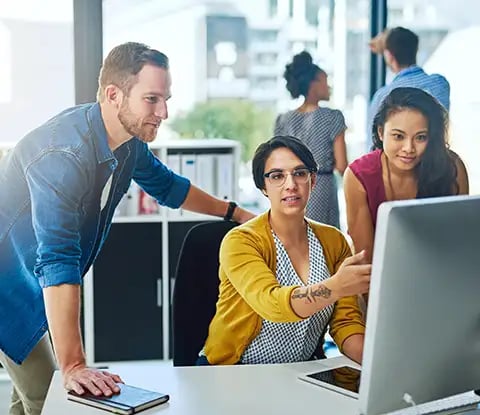 Three people are gathered around a computer screen in a modern office, working together, while two others collaborate in the background.